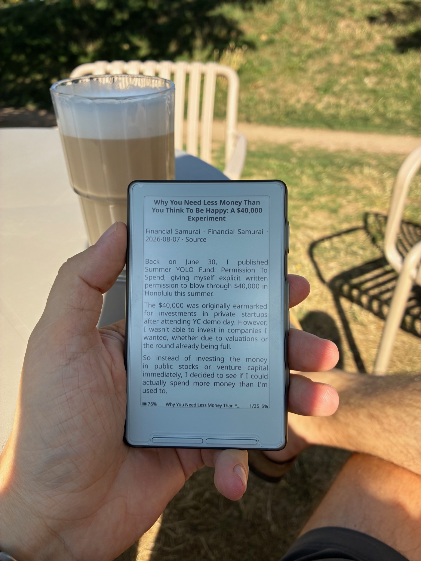 A hand holding the small X4 e-reader outdoors, its e-ink screen showing a blog post. A latte and a metal café chair sit in the sunlit background.